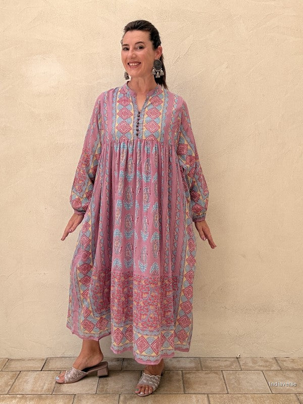 A woman modeling a long-sleeved, loose-fitting maxi dress with a pink, blue, and yellow bohemian-style geometric pattern, standing against a textured beige stucco wall.