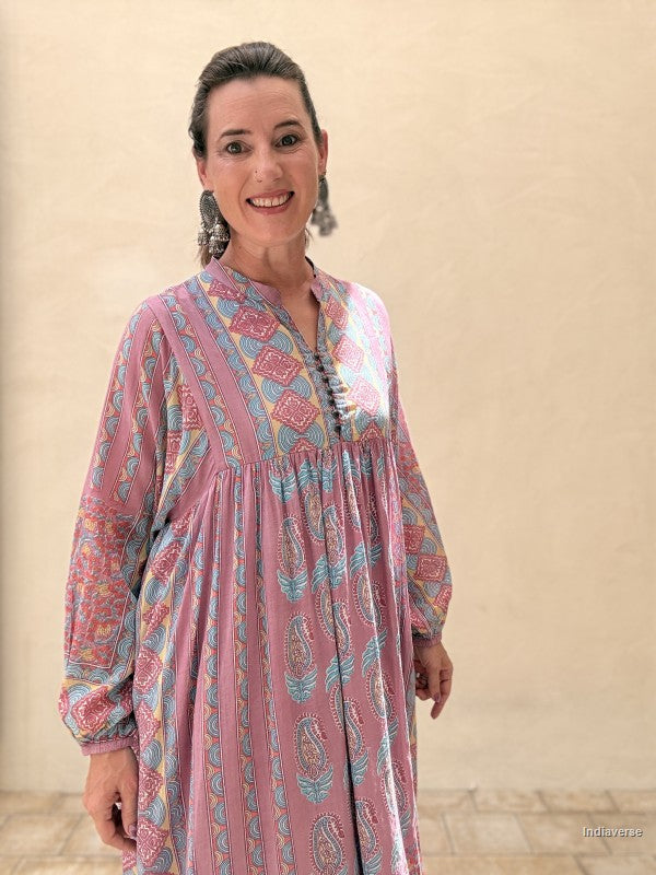 A woman wearing a pink long-sleeve bohemian tunic dress with blue and yellow paisley patterns, standing against a beige stucco wall.
