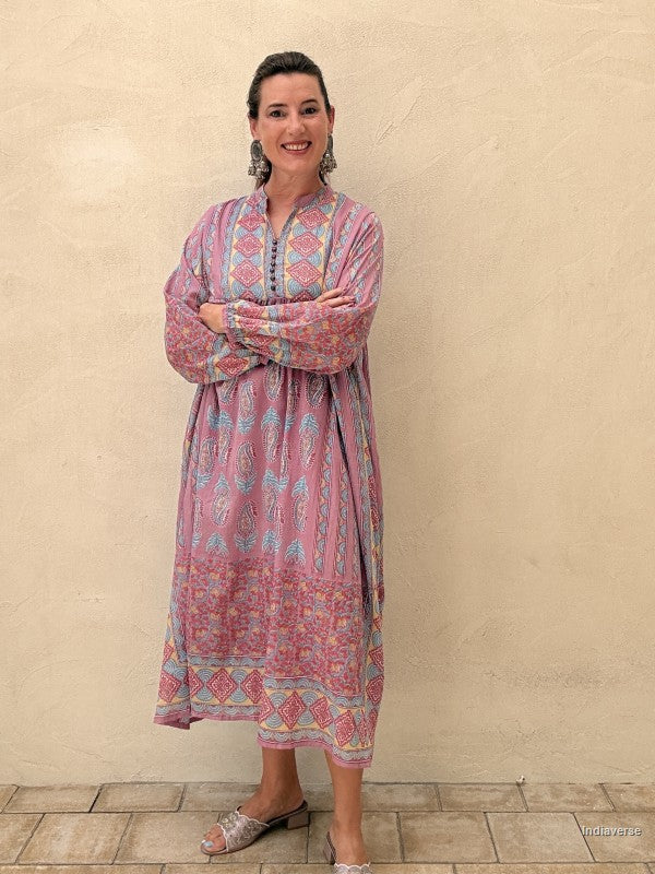 A woman modeling a long-sleeved, midi-length kaftan dress with a pink base and multicolor geometric patterns, standing against a textured beige stucco wall.