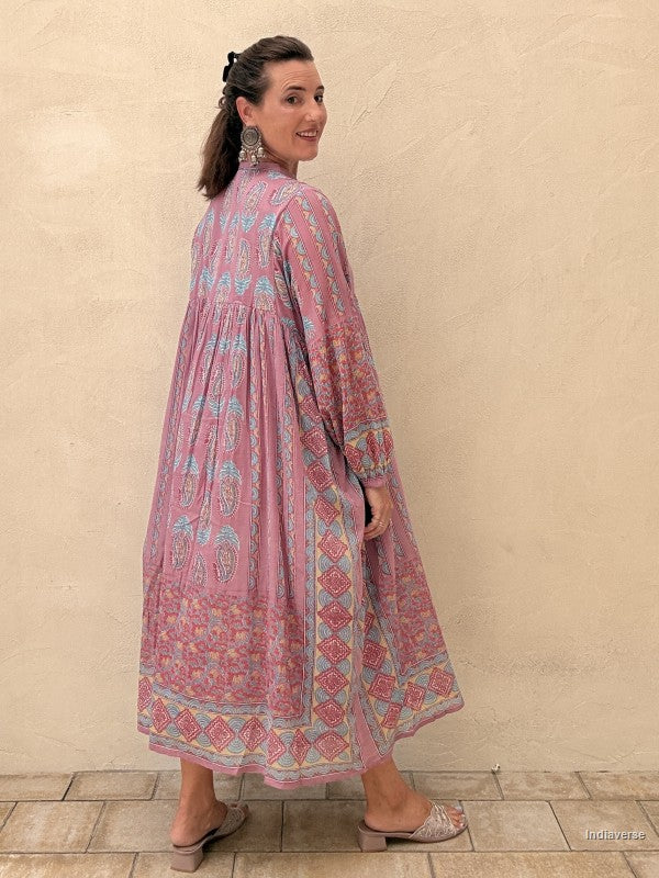 A woman modeling a long-sleeved, high-neck maxi dress with a pink and light blue paisley block print, standing against a textured beige stucco wall.