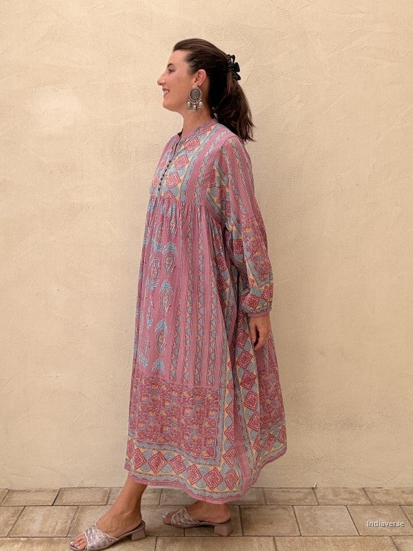 A woman wearing a pink and blue patterned long kaftan dress, standing in profile against a textured beige stucco wall.
