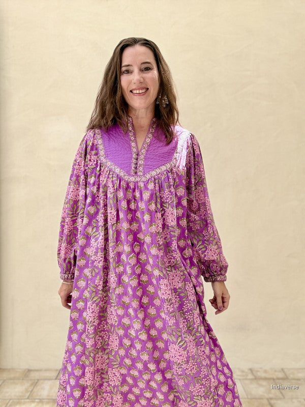 A woman wearing a purple and pink floral long-sleeve tunic dress, standing against a textured beige wall.