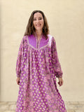 A woman wearing a purple and pink floral long-sleeve tunic dress, standing against a textured beige wall.