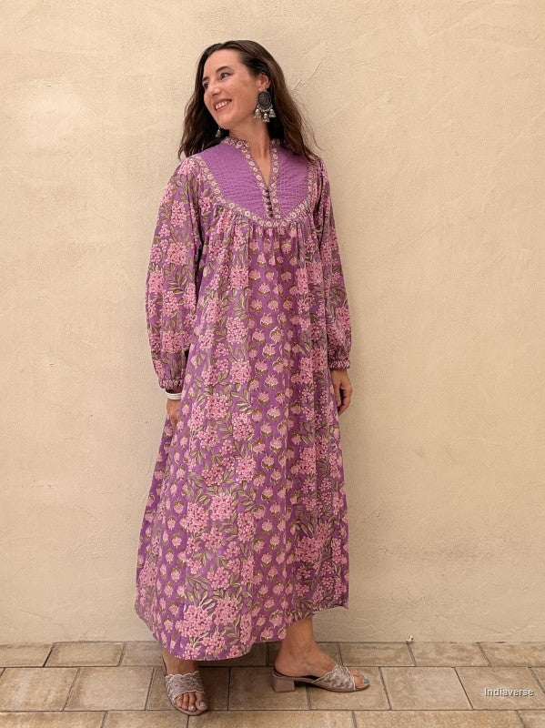 A woman wearing a purple and pink floral long-sleeve maxi dress with a matching purple embroidered yoke, standing against a textured beige stucco wall.