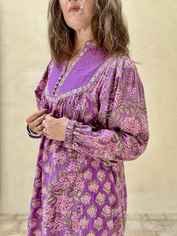 A woman wearing a purple long-sleeve tunic dress with a pink and green floral pattern, standing against a neutral beige wall.