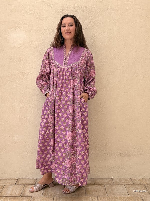 A woman wearing a long-sleeved, knee-length purple and pink floral print tunic dress with a purple collar, standing against a textured beige stucco wall.