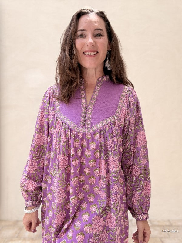 A woman wearing a purple and pink floral long-sleeve tunic dress, standing against a neutral beige wall.