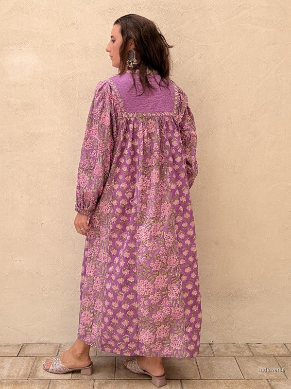 A woman wearing a long-sleeved purple and pink floral maxi dress, shown from the back against a textured beige wall.