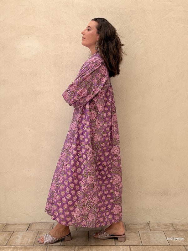 A woman wearing a long-sleeved, loose-fitting purple maxi dress with a pink and green floral pattern, standing against a textured beige stucco wall.
