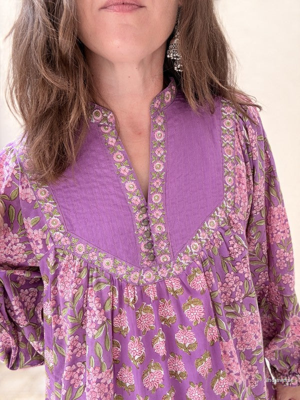 A close-up of a woman wearing a purple long-sleeve blouse with a pink and green floral print, featuring a gathered V-neckline with embroidered trim.