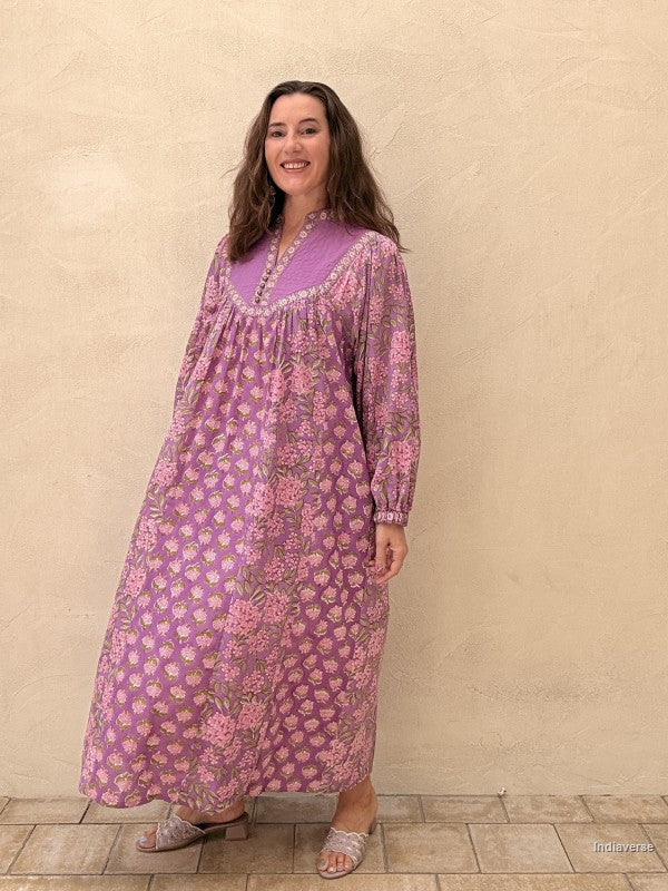 A woman modeling a long-sleeved, floor-length purple maxi dress with a pink and green floral pattern, standing against a textured beige wall.
