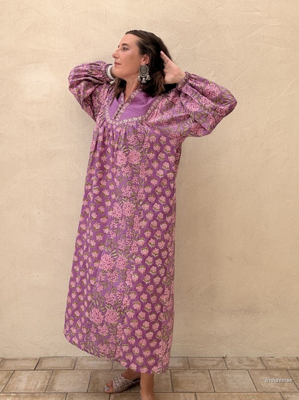 A woman wearing a long lavender-purple kaftan dress with a pink floral pattern, standing against a textured beige stucco wall.