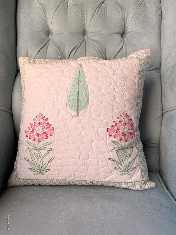 A square quilted throw pillow in pale blush pink with embroidered pink flowers and a mint green leaf, resting on a gray tufted armchair.