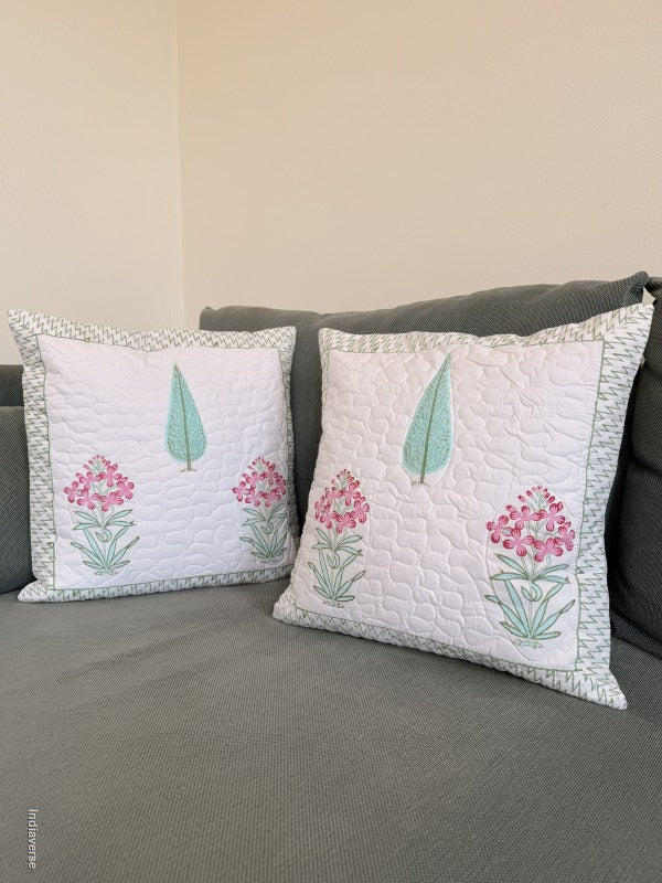 Two square decorative throw pillows with white quilted fabric, pink floral motifs, and mint green leaves, styled on a gray sofa against a beige wall.