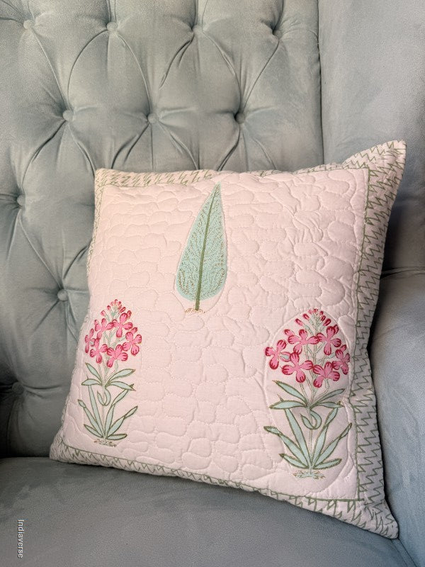 A square quilted throw pillow with a pale pink background, featuring embroidered pink flowers and a green leaf, resting on a gray tufted armchair.