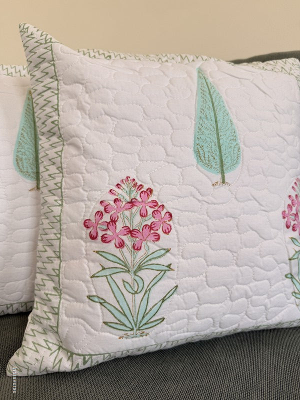 A close-up of a white quilted throw pillow featuring embroidered pink flowers and mint green leaves, resting on a dark gray textured sofa.