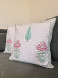 A white quilted throw pillow with embroidered pink flowers and a green leaf, styled on a dark gray sofa next to another matching pillow.