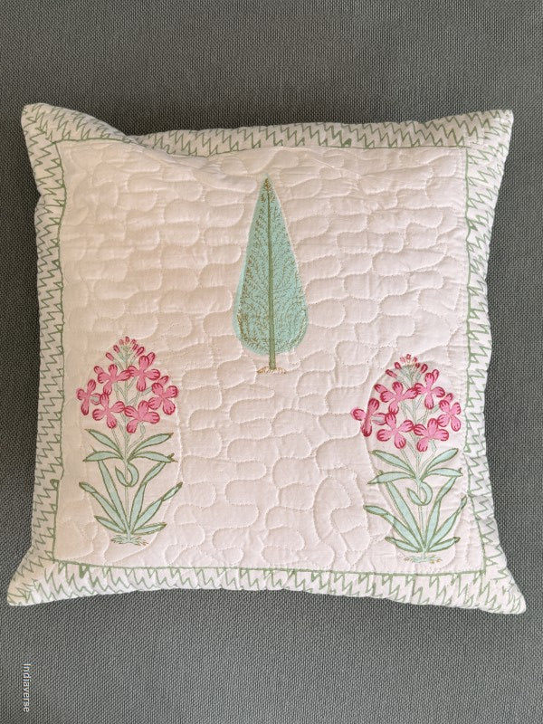 A square quilted throw pillow with a light pink center, featuring a green pine tree and two pink wildflower motifs, finished with a textured green and white patterned border, shown on a dark gray fabric background.