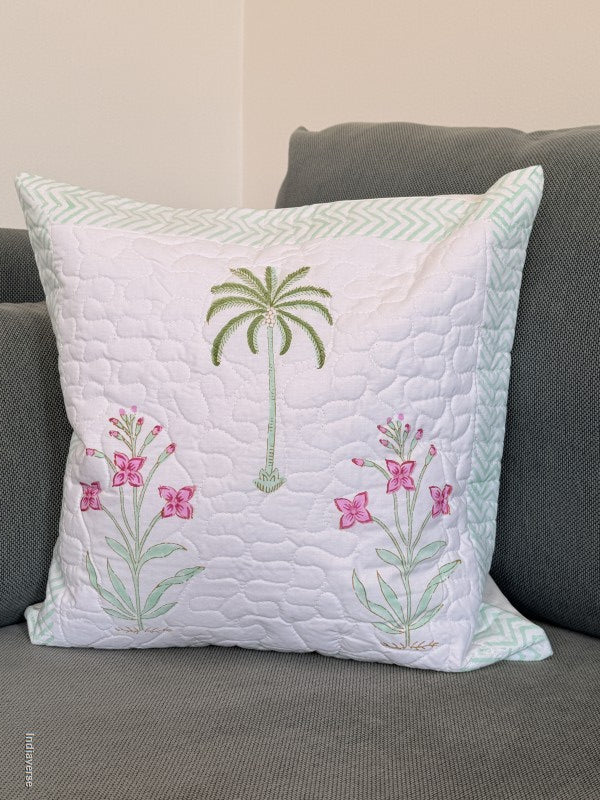 A square white quilted throw pillow with embroidered tropical designs, including a green palm tree and pink flowers, resting on a gray sofa.