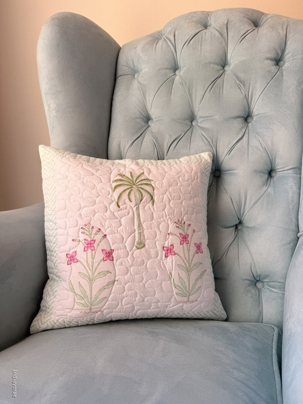 A square quilted throw pillow with a pink tropical design featuring a palm tree and pink flowers, resting on a light blue tufted armchair.