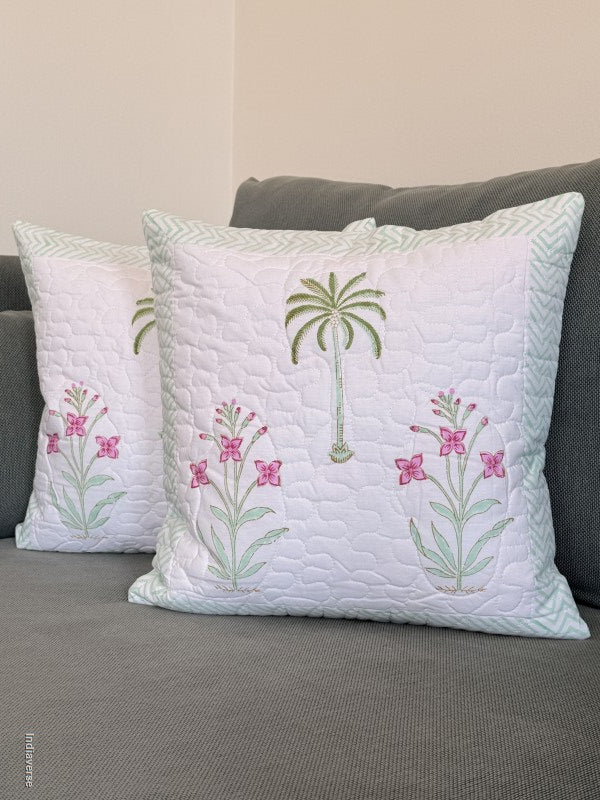 Two white quilted throw pillows with embroidered green palm trees and pink flowers, resting on a gray sofa against a beige wall.