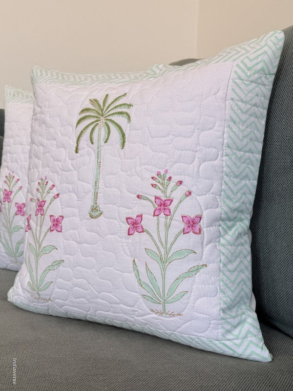 A square quilted throw pillow with a white center, pink floral embroidery, a green palm tree motif, and a mint green chevron border, resting on a gray sofa.