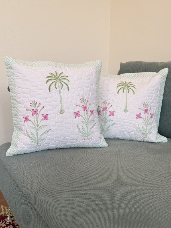 Two square white throw pillows with embroidered green palm trees and pink flowers, resting on a gray sofa against a beige wall.