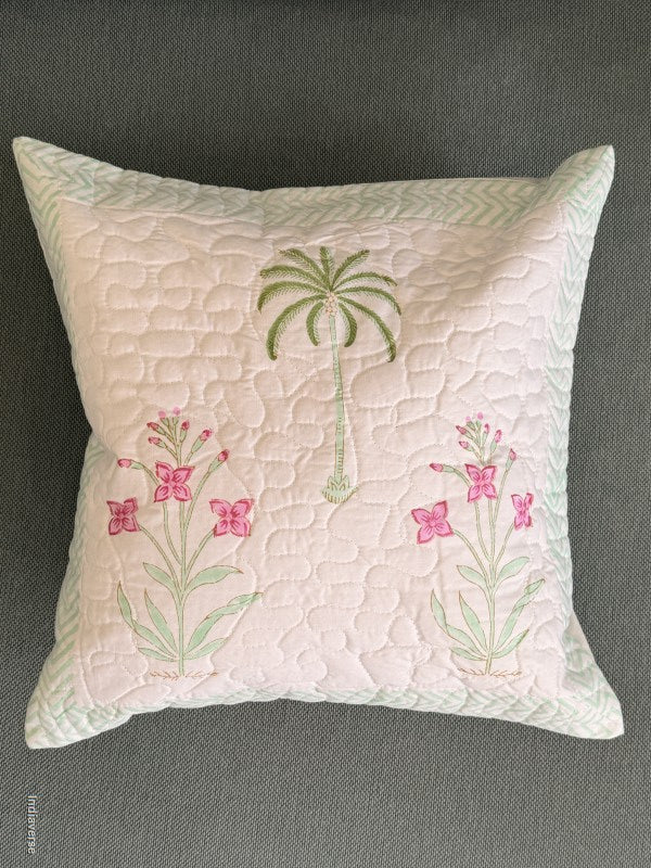 A square quilted throw pillow in pale pink with embroidered green palm trees and pink flowers, set against a dark gray textured background.