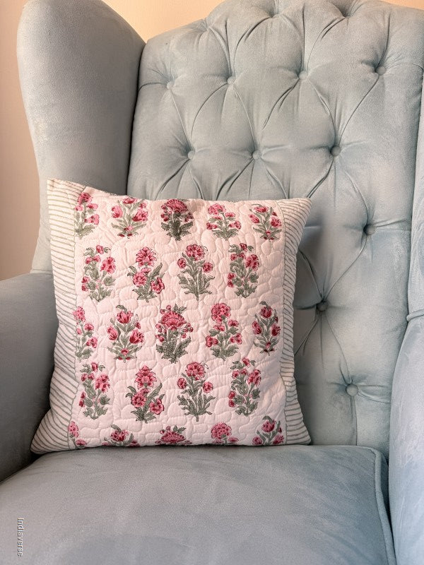A square quilted throw pillow with a pink and green floral pattern on a white background, featuring a striped green and white border, resting on a gray sofa.