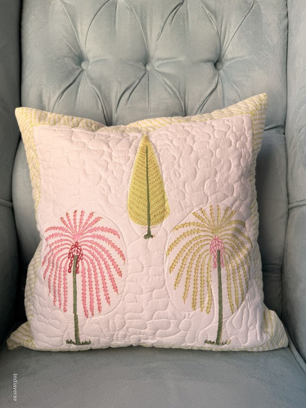 A square quilted throw pillow with a pale pink background, featuring embroidered palm trees in pink, yellow, and green, resting on a gray tufted armchair.