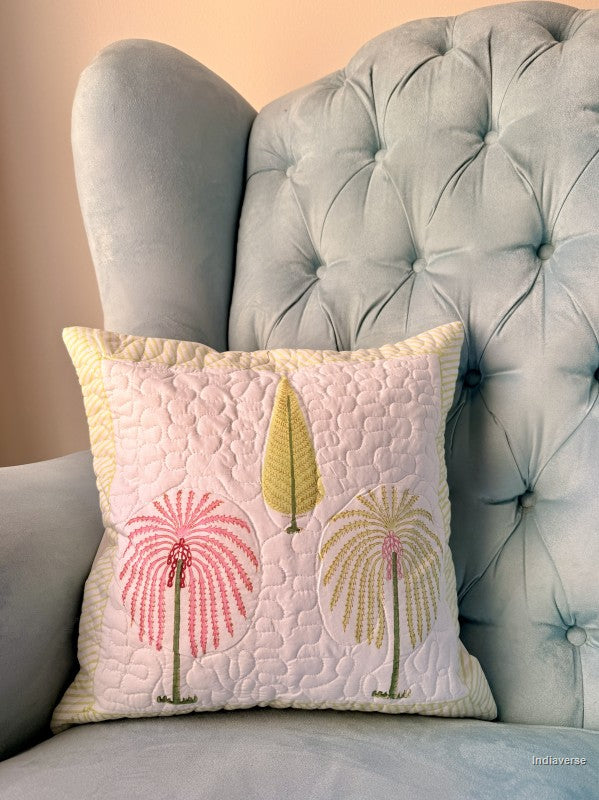 A square quilted throw pillow with a pink and yellow palm tree design, resting on a light blue tufted armchair.