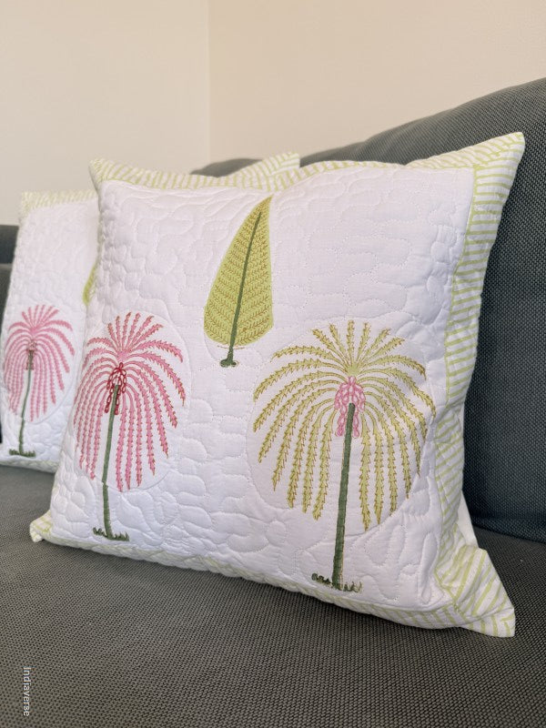 A white quilted throw pillow with embroidered palm trees in pink and olive green, resting on a dark gray sofa next to another matching pillow.