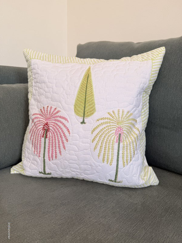 A square white quilted throw pillow with embroidered palm trees in pink, yellow-green, and green, resting on a gray fabric sofa.