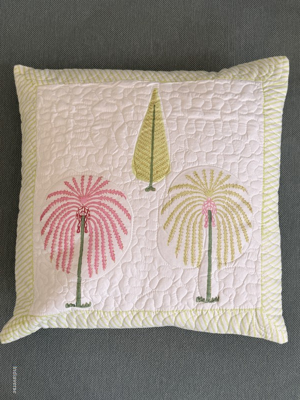 A square quilted throw pillow featuring embroidered palm trees in pink, green, and yellow on a pale pink background, finished with a striped yellow and white border, shown on a dark gray textured fabric background.