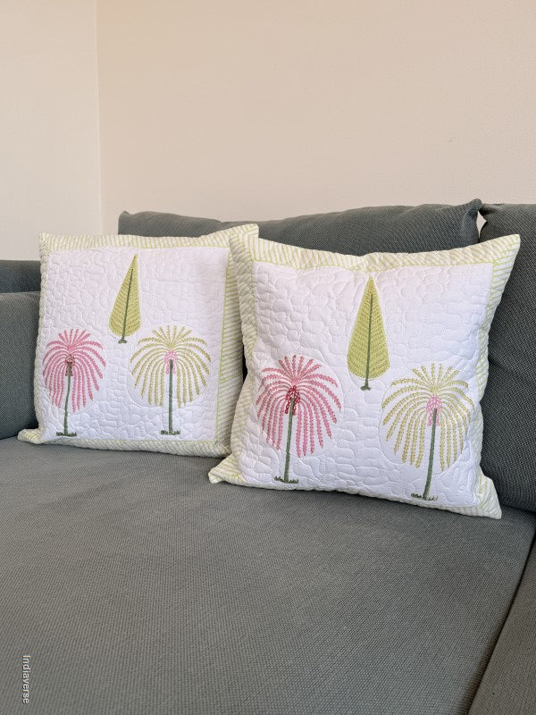 Two square white quilted throw pillows with embroidered palm tree designs in pink and green, resting on a gray fabric sofa against a beige wall.