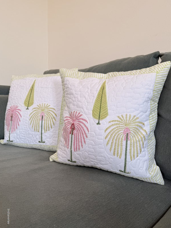 Two square decorative throw pillows with a white quilted background and embroidered palm tree motifs in pink and olive green, resting on a gray fabric sofa against a beige wall.