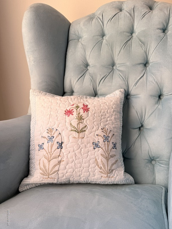 A square quilted throw pillow with embroidered floral and butterfly motifs in beige, blue, and pink, resting on a light blue tufted wingback armchair.