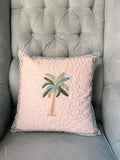 A square quilted throw pillow with a palm tree design, resting on a tufted gray armchair. The pillow is light pink with a blue and green patterned border and features an embroidered palm tree motif in the center.