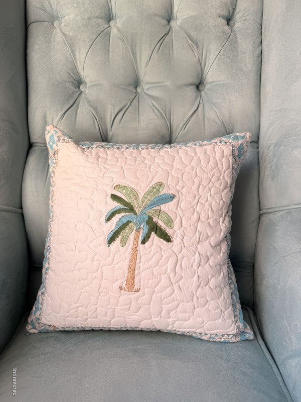 A square quilted throw pillow with a palm tree design, resting on a tufted gray armchair. The pillow is light pink with a blue and green patterned border and features an embroidered palm tree motif in the center.