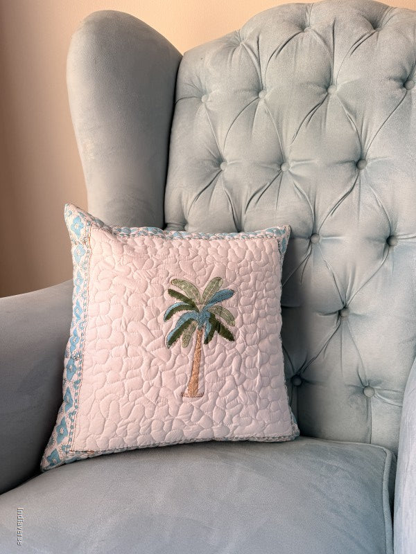 A square decorative throw pillow with a light pink quilted body, a central embroidered palm tree, and a blue patterned border, resting on a light blue tufted armchair.