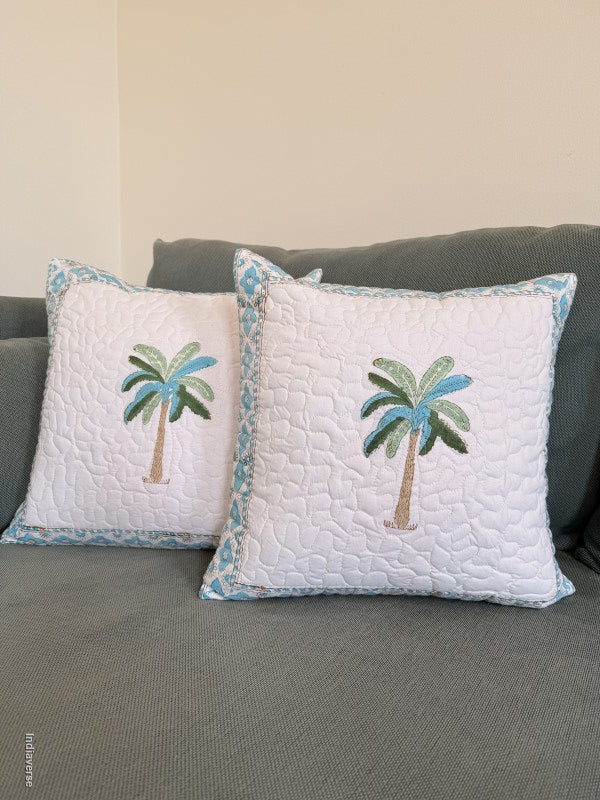 Two white quilted throw pillows with embroidered palm trees and blue patterned borders, displayed on a gray sofa against a beige wall.