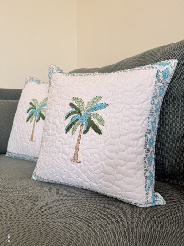 A white quilted throw pillow with an embroidered palm tree design sits on a dark gray sofa, with a second matching pillow partially visible behind it.