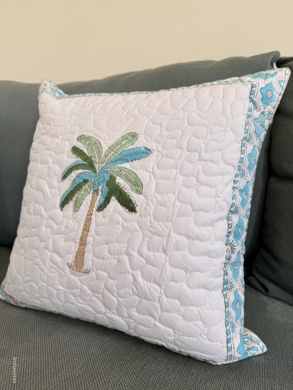 A square white quilted throw pillow with an embroidered palm tree design and a light blue patterned border, resting on a gray sofa.