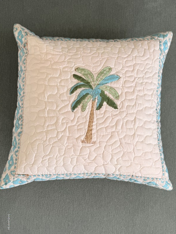 A square decorative throw pillow with a quilted cream center, a light blue patterned border, and an embroidered palm tree motif in green, blue, and tan on the front, shown against a dark gray textured background.