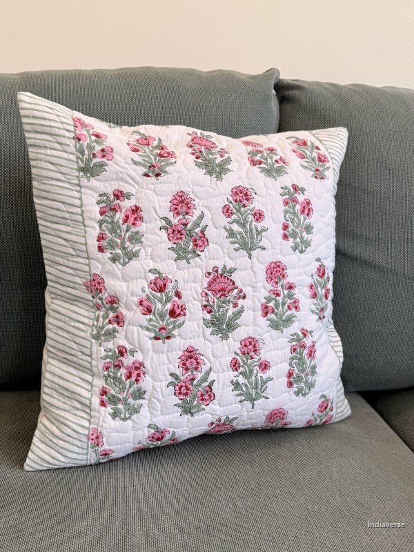 A square decorative throw pillow with a pink and green floral quilted pattern and striped fabric borders, resting on a gray fabric sofa.