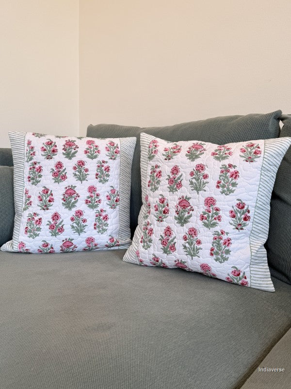 Two square decorative throw pillows with a pink and green floral block print on a white background, resting on a gray fabric sofa against a beige wall.