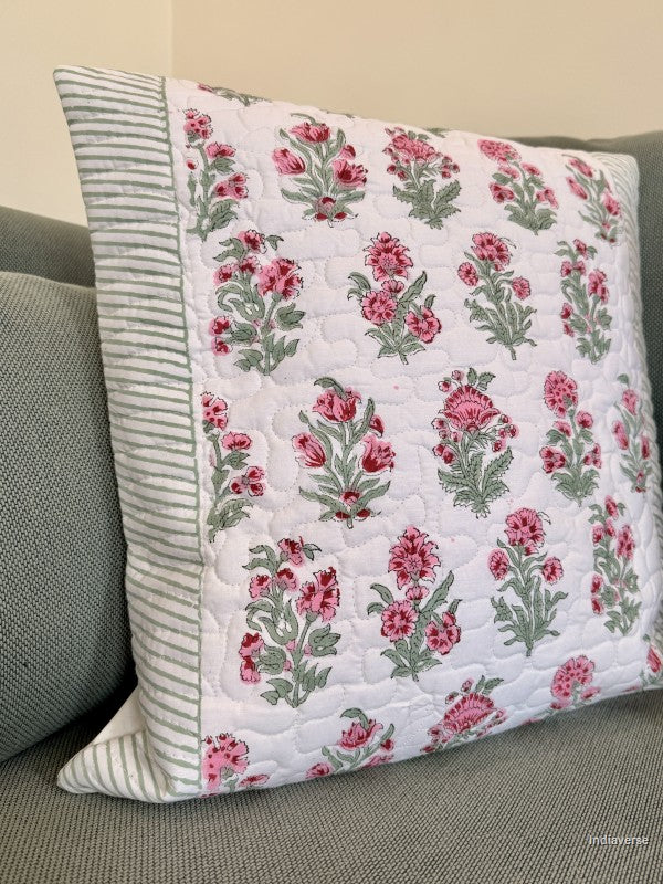 A square quilted throw pillow with a pink and green floral pattern on a white background, featuring a striped green and white border, resting on a gray sofa.