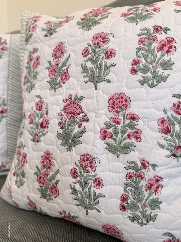 A close-up of a quilted throw pillow featuring a pink and green floral pattern on a white background, resting on a gray sofa.