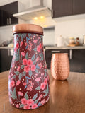 Decorate water jar made from copper with red and pink floral design sitting on a brown surface in a kitchen with a copper cup in the background