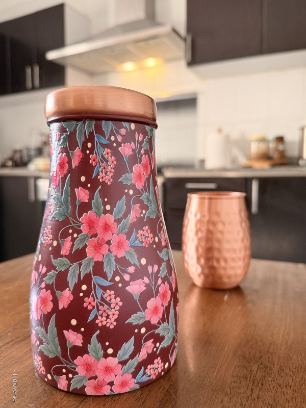 Decorate water jar made from copper with red and pink floral design sitting on a brown surface in a kitchen with a copper cup in the background